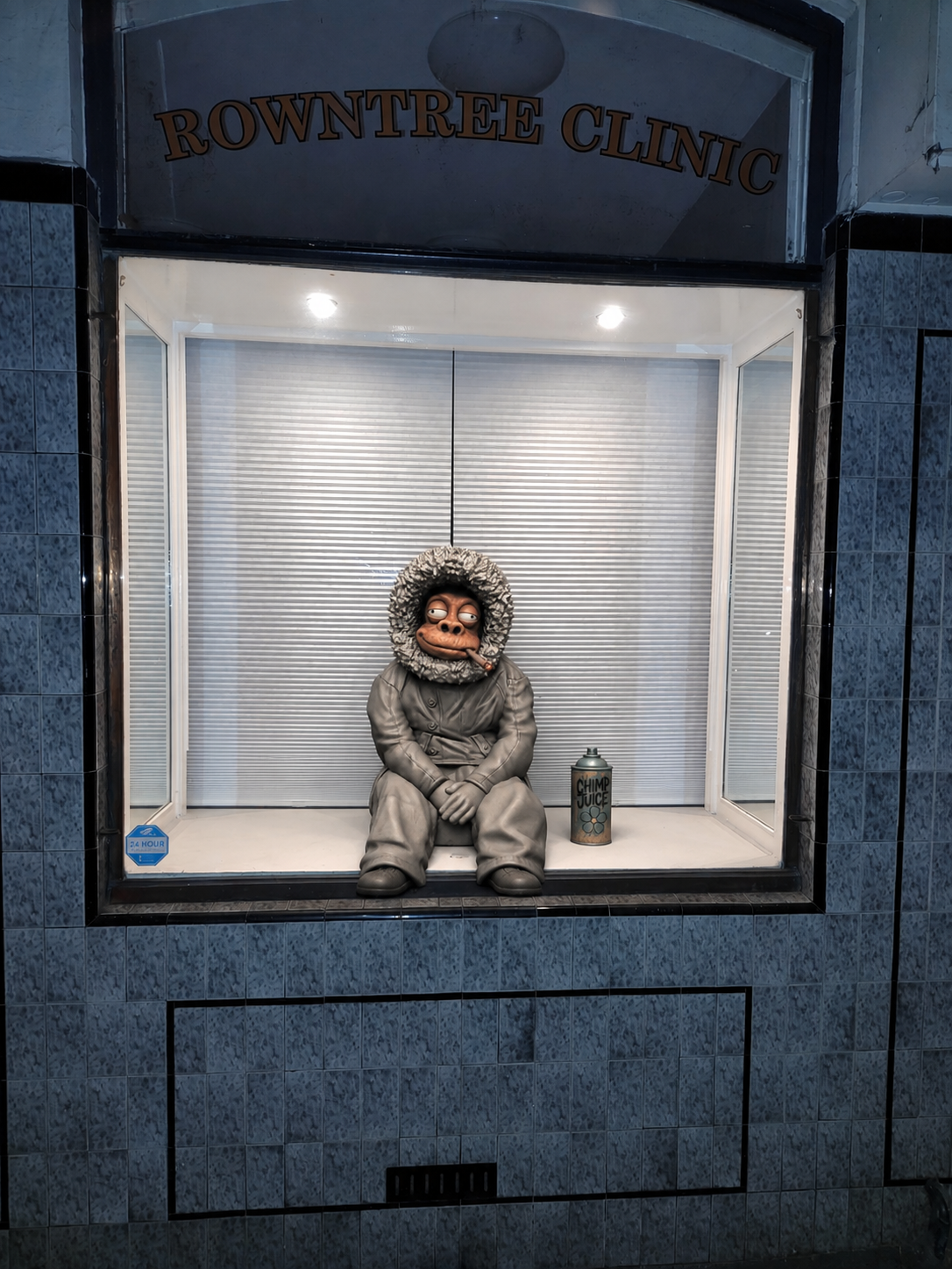 A grey sculpted chimpanzee in a hooded coat sits in the display window of a former Balmain clinic, beside a spray can, waiting.